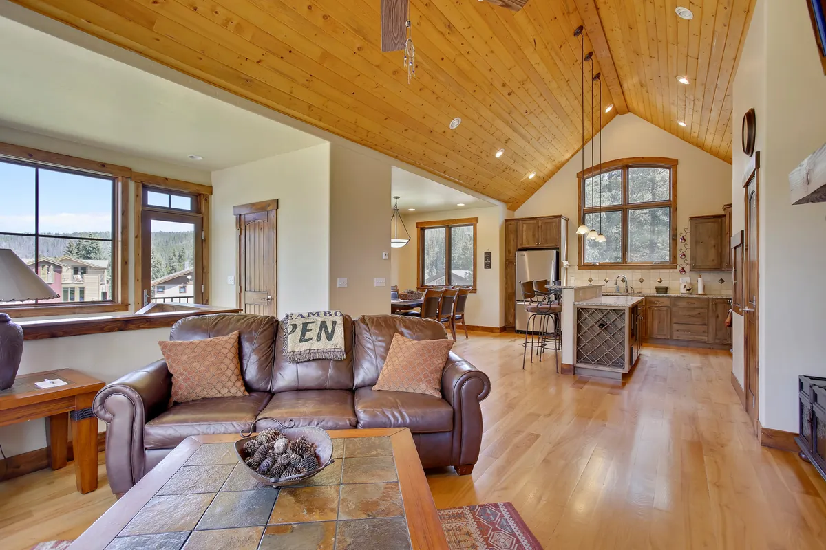 A comfortable living area featuring a plush leather sofa, a warm wooden ceiling, and expansive windows that flood the space with natural light. The kitchen area is visible in the background, indicating a spacious, open-concept design ideal for groups or families.