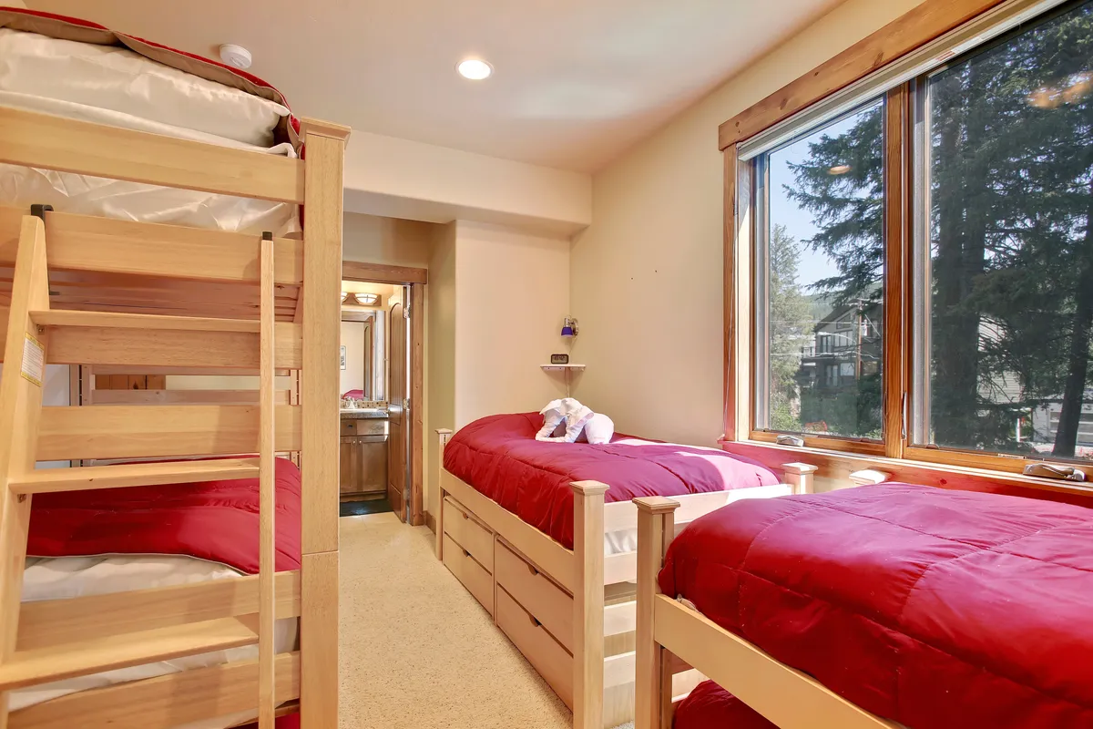 A cheerful bunk room featuring a sturdy wooden bunk bed and a twin bed with bright red bedding. Sunlight streams through the window, creating a warm, playful environment ideal for families or groups. The layout offers flexibility and comfort for multiple guests.