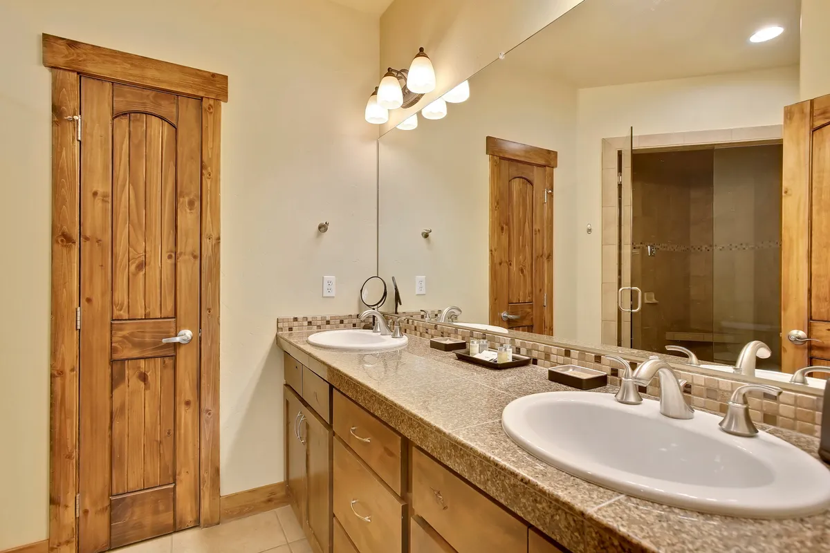 A well-appointed bathroom with double vanity sinks, wooden cabinetry, a large framed mirror, and a glimpse of the shower area. The space is bright, clean, and designed for efficient morning and evening routines, offering both privacy and comfort.