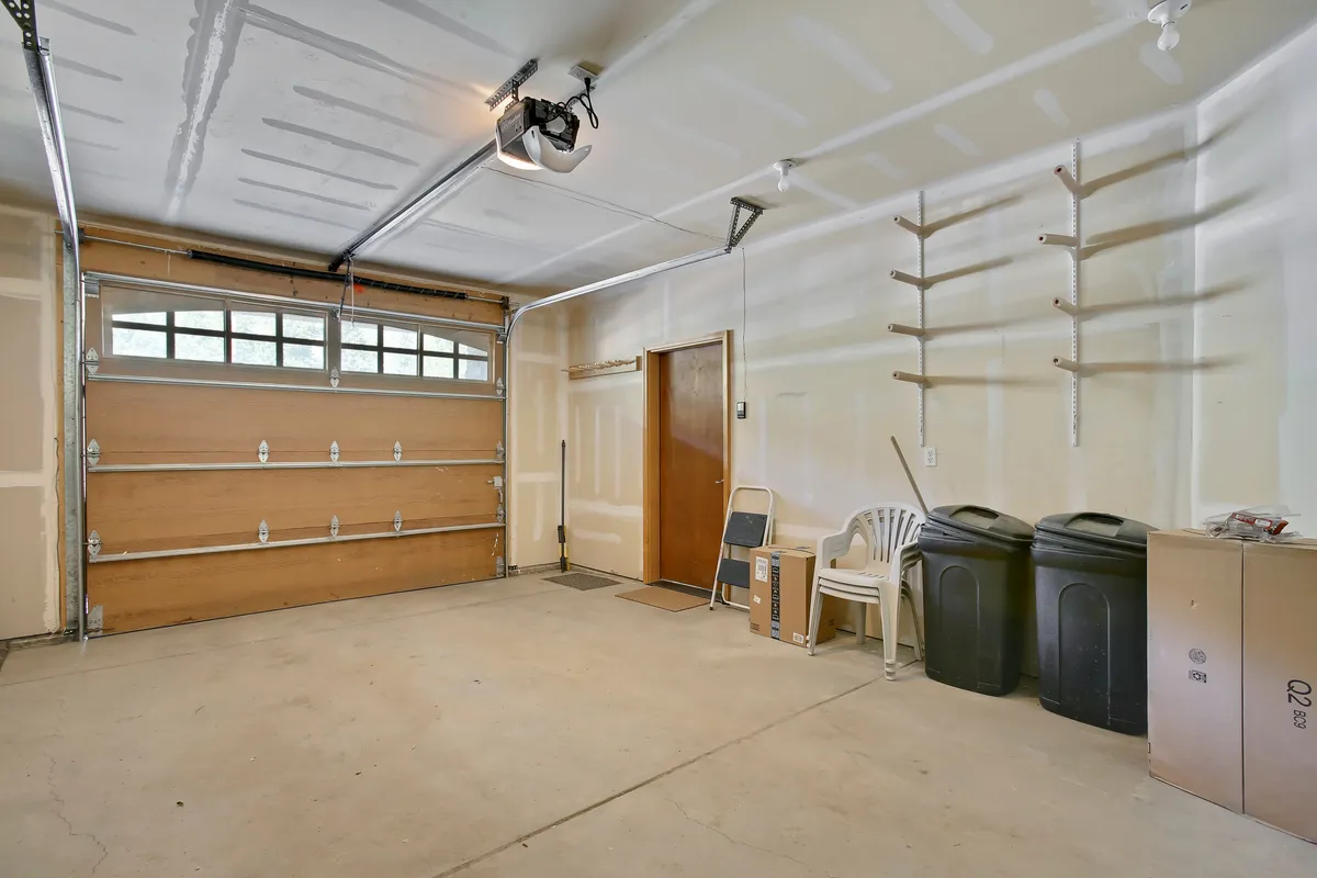 A garage interior with a visible garage door, storage shelves, cardboard boxes, trash bins, and a chair. The space is practical and organized, showing capacity for storage and utility.