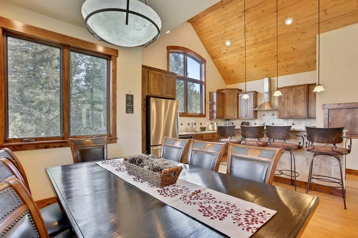 This image features a spacious dining table set near large windows, with a wooden ceiling and a kitchen island visible in the background. The setting is bright and welcoming, with a harmonious blend of kitchen and dining spaces that invite both family gatherings and festive meals.