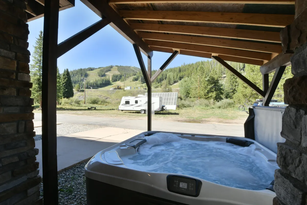An inviting outdoor hot tub set against a dramatic mountain backdrop, with rustic wooden beams and a stone structure. The serene scene suggests relaxation and connection to nature after a day of adventure.