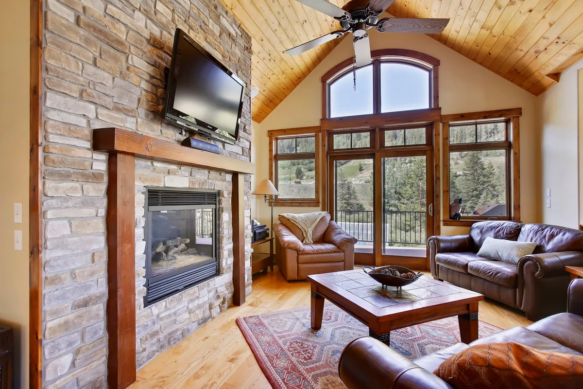 A spacious great room adorned with leather sofas arranged around a striking stone fireplace. Large windows allow natural light to flood the room, offering breathtaking mountain views. A wooden ceiling adds warmth and character, while a coffee table anchors the seating area, ideal for board games or shared snacks.